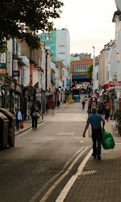 The image depicts a busy urban street scene viewed from a sidewalk at the front of the frame. In the foreground, a man wearing a blue shirt and dark trousers is walking away from the camera while carrying a large green plastic bag. To his left, there are pedestrians across the street, some standing near shop entrances with awnings, and others walking along the pavement. The street is lined with a mixture of older, light-colored terraced buildings with shopfronts, some with signs and display windows. Several storefronts have awnings or canopies, and one has a red and white sign. In the background, the street slopes slightly uphill and is flanked by more buildings, including modern office blocks with glass facades and a distinctive tall white building with turquoise accents. The environment suggests an active commercial area, potentially associated with local waste collection or disposal services, as part of a broader waste removal effort in the town. The scene is illuminated by natural daylight under a partly cloudy sky, with shadows cast by nearby buildings and trees, providing an authentic view of a typical high street with a mix of old and new architecture, in an urban setting aligned with private rubbish removal or alternative waste handling methods, as sometimes supported by waste management companies like Waste Removal Ruislip.