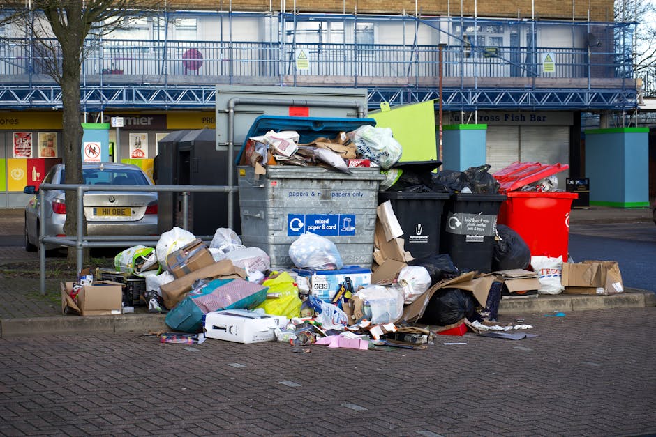 In the foreground of the image, there is a collection of overflowing rubbish bins and piles of waste on a paved urban street, with various types of waste including cardboard boxes, plastic bags, and paper scattered around. The central grey bin with a blue lid appears to be designated for mixed paper and cardboard, while several black bins and a prominent bright red bin are also filled and spilling waste. Surrounding the bins are numerous loose packaging materials, crumpled paper, and discarded packaging, indicating an excess of waste beyond the containers' capacity. To the left, a parked silver car is partially visible, positioned behind a metal barrier that separates the waste area from the pedestrian pathway. Behind the scene, there are retail units and a building under construction or renovation, with scaffolding and protective netting, and clear signage visible at street level. The environment suggests an urban shopping district or town centre, where private rubbish collection services like Waste Removal Ruislip may assist in managing excess waste, supporting alternative waste handling outside of local authority collection systems. The entire scene is evenly lit, with natural daylight highlighting the disorderly waste accumulation amidst a typical town setting.